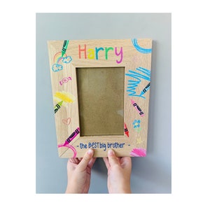 Puede incluir: Un marco de fotos de madera con un diseño de crayones de colores. El marco tiene un espacio para una foto y el texto "Harry - the BEST big brother -" está escrito en la parte inferior.