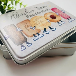 May include: A silver metal tin with a white lid featuring a cartoon illustration of various biscuits with legs and arms. The text on the lid reads "Aleeha's team Biscuits stash".