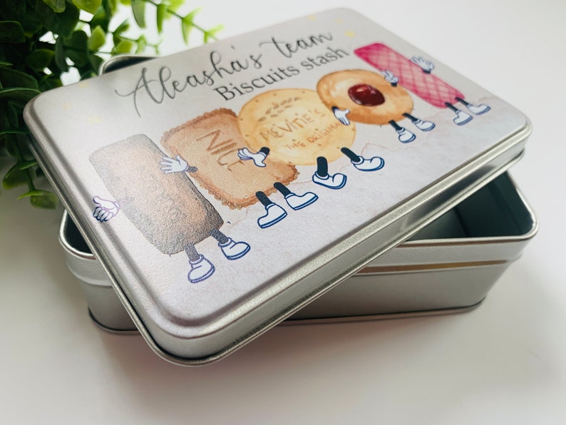 May include: A silver metal tin with a white lid featuring a cartoon illustration of various biscuits with legs and arms. The text on the lid reads "Aleeha's team Biscuits stash".