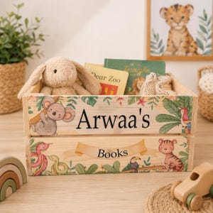 Op de afbeelding: Houten opbergkrat met een junglethema, met een illustratie van een koala, slang en tijger. De krat is gepersonaliseerd met de naam "Arwaa's" en het woord "Books". Binnenin liggen boeken en een knuffelkonijn.