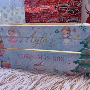 Puede incluir: Una caja de madera azul con temática navideña. La caja tiene un fondo blanco con una inscripción dorada que dice "Ayla's Christmas Box". La caja está decorada con imágenes de un cascanueces, una bailarina, un caballito de balancín y un árbol de Navidad.