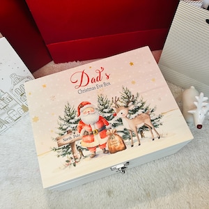 Puede incluir: Una caja rectangular de madera con tapa abatible que presenta una ilustración impresa de Santa Claus y un reno en un bosque nevado. El texto "Dad's Christmas Eve Box" está impreso en la tapa, junto con un letrero de "North Pole" y copos.