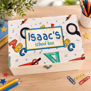 May include: A wooden school box with a light wood finish. The lid is decorated with colourful illustrations of school supplies, ABCs, and the text "Isaac's school box" in a blue and yellow frame. A metal clasp secures the box.