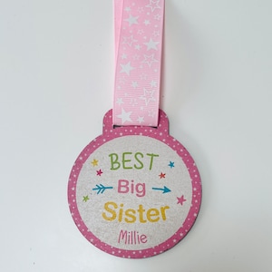 May include: Pink and white wooden medal with the text "BEST Big Sister Millie" in colourful letters and arrows. The medal is attached to a pink ribbon with white stars.