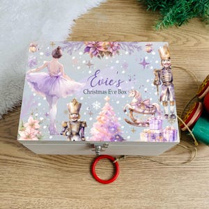 Puede incluir: Una caja rectangular de madera blanca con una ilustración festiva de una bailarina con tutú morado, dos cascanueces, un caballito balancín, un árbol de Navidad y regalos. El texto dice Evie's Christmas Eve Box en letra morada.