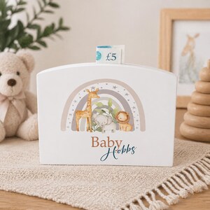 Könnte beinhalten: Weiße Baby-Spardose aus Holz mit Regenbogen- und Safari-Tiermotiv. Die Box zeigt eine Illustration von Giraffe, Elefant und Löwe. Der Text "Baby Hobbs" steht darunter. Eine Fünf-Pfund-Note steckt im Schlitz.