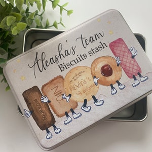 May include: A white metal tin with a light brown background and a colourful illustration of cartoon biscuits. The tin has the text "Aleasha's team Biscuits stash" printed on it.