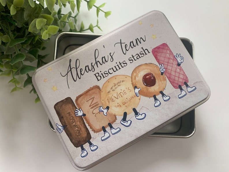 May include: A white metal tin with a light brown background and a colourful illustration of cartoon biscuits. The tin has the text "Aleasha's team Biscuits stash" printed on it.