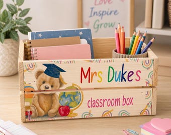 May include: Wooden classroom box featuring a teddy bear in a graduation cap, a globe, and an apple. The box is personalised with the name "Mrs Dukes" and the words "classroom box" in colourful letters. Contains notebooks and pencils.
