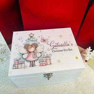 Puede incluir: Una caja blanca de Nochebuena con una ilustración decorativa de un hada, un árbol de Navidad, regalos y mariposas. La caja está personalizada con el nombre "Gabriella's" y el texto "Christmas Eve Box". Tiene un cierre plateado.