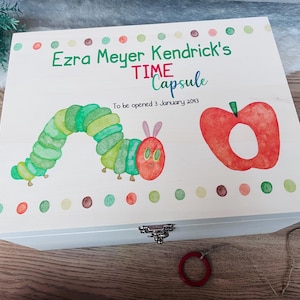 May include: A white wooden time capsule box with a watercolor illustration of a green caterpillar and a red apple. The box is decorated with colorful dots and the text "Ezra Meyer Kendrick's TIME Capsule".