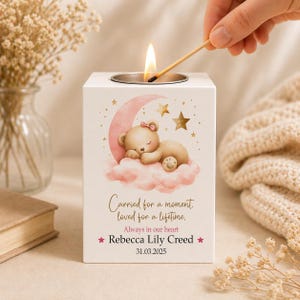 May include: A white candle holder with a metal top, decorated with a sleeping teddy bear on a pink cloud and moon. The text reads "Carried for a moment, loved for a lifetime, Always in our heart, Rebecca Lily Creed, 31.03.2025". A match is lit above the candle.