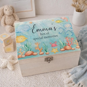 Könnte beinhalten: Eine Holzkiste mit hellblauem Deckel, verziert mit einer Unterwasserszene mit Cartoon-Fischen, Korallen und dem Text "Emma's box of special memories". Die Kiste hat einen silbernen Verschluss.