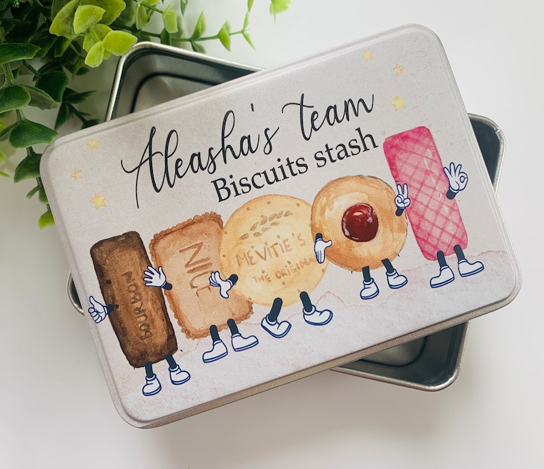 May include: A white metal tin with a lid, featuring a cartoon illustration of various biscuits with arms and legs. The tin is decorated with gold stars and the text "Aleasha's team Biscuits stash".