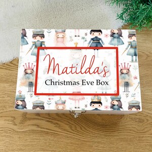 Puede incluir: Caja de madera blanca con tapa con bisagras y cierre de plata. La caja está decorada con un patrón de ilustraciones de acuarela de soldados cascanueces y bailarinas. El texto "Matilda's Christmas Eve Box" está impreso en la parte delantera de la caja.