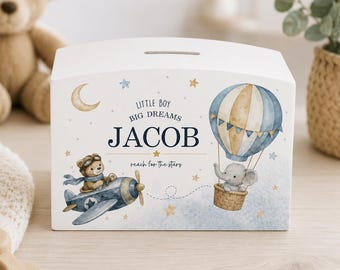 May include: A white wooden money box featuring a slot on top and watercolour illustrations of a bear in a blue aeroplane and an elephant in a hot air balloon. The text reads LITTLE BOY BIG DREAMS JACOB reach for the stars with gold stars.