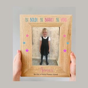Puede incluir: Un marco de fotos de madera con una foto de una niña con un vestido negro. El marco tiene las palabras "BE BOLD! BE BRAVE! BE YOU!" en la parte superior y "Annie's first day at Siddal Primary School" en la parte inferior. Hay estrellas rosas y azules alrededor de la foto.