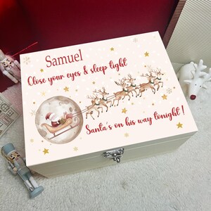Puede incluir: Una caja de recuerdos rectangular de madera blanca con un diseño impreso de Santa en un trineo tirado por renos, una luna llena, estrellas doradas y el texto: Samuel, Close your eyes & sleep tight, Santa's on his way tonight! Tiene un cierre metálico.