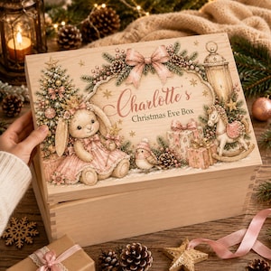 Puede incluir: Una caja rectangular de madera con una ilustración impresa de un conejo con vestido rosa, un caballo balancín, un pájaro y un árbol de Navidad. El texto "Charlotte's Christmas Eve Box" está impreso en el centro, enmarcado por un lazo rosa.