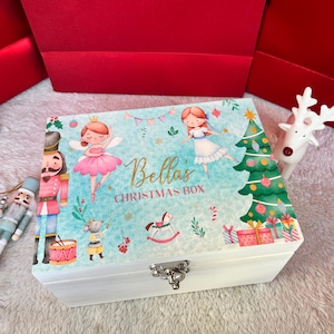 Puede incluir: Una caja rectangular de madera blanca con tapa abatible que presenta un fondo azul claro e ilustraciones coloridas de un cascanueces, una bailarina, un ratón, un caballito de madera y un árbol de Navidad. El texto dice Bella's CHRISTMAS BOX.