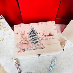 Puede incluir: Una caja de madera rectangular para Nochebuena con base blanca y tapa clara que presenta una ilustración impresa de un conejo con tutú rosa junto a un árbol de Navidad y regalos, con el texto Sophie Christmas Eve Box.