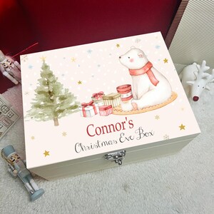 Puede incluir: Una caja rectangular de madera con tapa abatible que presenta una ilustración en acuarela de un oso polar con bufanda roja, un árbol de Navidad verde y regalos. El texto "Connor's Christmas Eve Box" está impreso en rojo y negro.