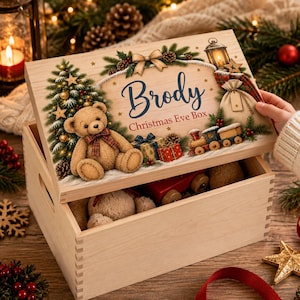 Puede incluir: Una caja de madera clara con una tapa que presenta una ilustración impresa de un oso de peluche, un árbol de Navidad, un tren de juguete y un regalo. La tapa muestra el texto Brody Christmas Eve Box en letras azules y rojas.