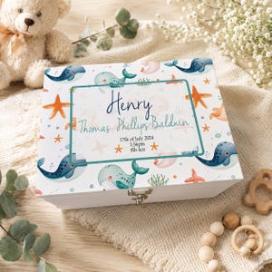 Puede incluir: Caja de recuerdos de madera blanca decorada con ballenas, estrellas de mar y algas marinas. La tapa de la caja presenta el nombre "Henry" y los detalles del nacimiento: fecha, hora y peso (3,74 kg). La caja tiene un cierre metálico.