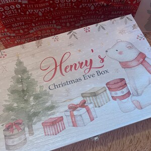 Puede incluir: Una caja rectangular de madera con acabado blanco que presenta una ilustración de un oso polar con bufanda roja, un árbol de Navidad verde y varios regalos envueltos. El texto Henry's Christmas Eve Box está impreso en rojo y negro.