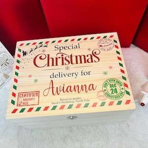 Puede incluir: Una caja rectangular de madera con tapa abatible que presenta rayas diagonales rojas y verdes en los bordes. La tapa está impresa con el texto Special Christmas delivery for Avianna, Santa's Workshop, North Pole y varios sellos navideños.