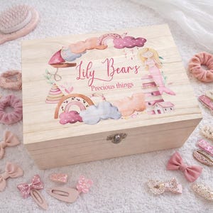 Può includere: Una scatola di legno color chiaro con coperchio incernierato, con un disegno ad acquerello di una sirena, arcobaleni e nuvole. Il testo "Lily Bears Precious things" è stampato sul coperchio. Vari accessori per capelli rosa circondano la scatola.