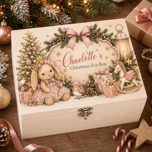 Puede incluir: Una caja rectangular de madera con cierre metálico, que presenta una ilustración impresa de un conejo con vestido rosa, un pájaro, un caballito de madera, regalos y un farol. El texto dice Charlotte's Christmas Eve Box, rodeado de ramas.
