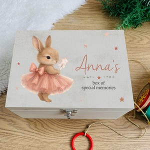May include: A light gray wooden box with a hinged lid. The lid features a watercolor illustration of a brown bunny wearing a pink dress and holding a star wand. The text "Anna's box of special memories" is written on the lid.