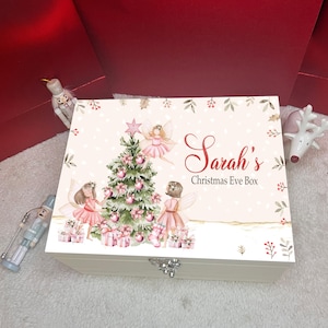 Puede incluir: Una caja rectangular de madera blanca con una ilustración navideña de un árbol decorado, dos hadas con vestidos rosas y cajas de regalo. El texto "Sarah's Christmas Eve Box" está impreso en letras rojas y negras en la tapa.