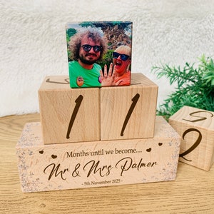 May include: Wooden block calendar featuring a photo of a couple. The calendar shows the numbers 11 and 2. The base reads "Months until we become... Mr & Mrs Palmer - 5th November 2025". A charming wedding countdown.