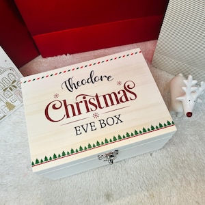 Puede incluir: Una caja rectangular de madera con tapa abatible y cierre de metal plateado. La tapa presenta el texto "Theodore Christmas EVE BOX" en letra negra y roja, acentuado por bordes decorativos verdes y rojos y pequeños motivos de pinos.