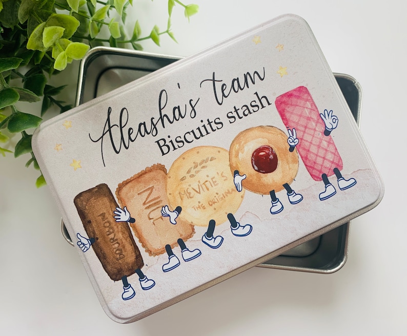 May include: A white metal tin with a colourful illustration of cartoon biscuits. The tin has the text "Aleasha's team Biscuits stash" on the front.