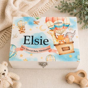 Op de afbeelding: Houten herinneringskist met een babythema. De kist toont een luchtballon met dieren en de naam "Elsie". De tekst "Very Special Baby Memories" is ook aanwezig. De kist is afgesloten met een zilveren sluiting.