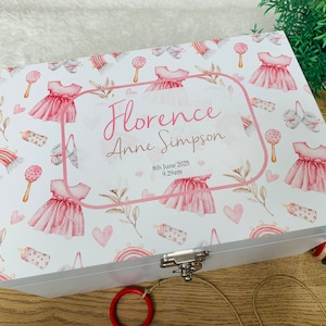 Peut inclure: Boîte à souvenirs en bois blanc avec un motif floral rose et blanc. La boîte a un couvercle à charnières avec une serrure et une clé. La boîte est décorée d'une robe rose, d'un hochet, d'un biberon et de cœurs. La boîte porte l'inscription "Florence Anne Simpson 8 juin 2025 9h29" sur le devant.