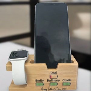 May include: A wooden phone and watch charging stand with a personalised message that reads "Dad, Emily, Bethany, Caleb, Happy Father's Day 2022". Each name has a battery icon next to it, indicating the charge level.