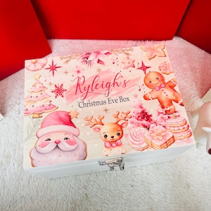 Puede incluir: Una caja rectangular de madera blanca con cierre metálico, con un estampado festivo de Papá Noel, un reno, un hombre de jengibre, árboles de Navidad y copos de nieve. El texto Ryleigh's Christmas Eve Box está impreso en el centro.