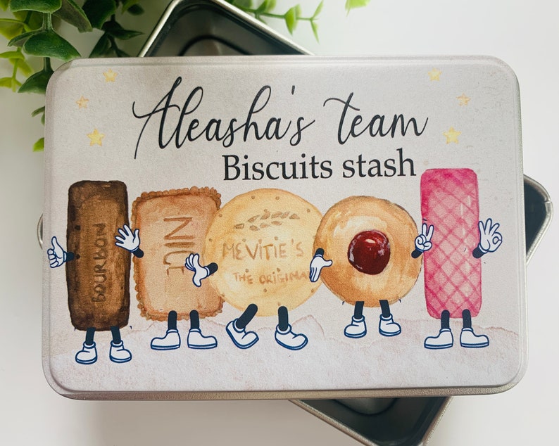 May include: A white metal tin with a colourful illustration of cartoon biscuits. The tin has the text "Aleasha's team Biscuits stash" on the front.