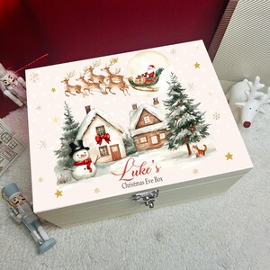 Puede incluir: Una caja rectangular de madera para Nochebuena con tapa abatible que presenta una escena invernal festiva. La ilustración incluye a Santa en un trineo con renos, casas cubiertas de nieve, un muñeco de nieve, un árbol de Navidad y el texto Luke's Christmas Eve Box.