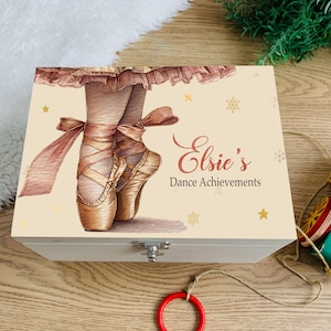 May include: A wooden box with a light beige lid featuring a watercolor illustration of ballet shoes. The box is decorated with gold stars and snowflakes, and the text "Elsie's Dance Achievements" is written in a cursive font.