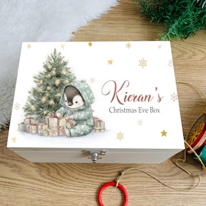 Puede incluir: Una caja rectangular de madera blanca con un gráfico de un pingüino con capucha sentado junto a un árbol de Navidad con regalos. El texto dice Kieran's Christmas Eve Box. La caja tiene un cierre de metal plateado y estrellas doradas.