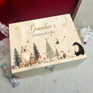 Puede incluir: Una caja rectangular de madera con tapa abatible y cierre de metal plateado. La tapa presenta el texto Brandan's Christmas Eve Box, rodeado de ilustraciones de osos, pinos y estrellas doradas sobre un fondo de color crema claro.