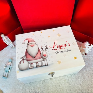 Puede incluir: Una caja rectangular de madera con tapa abatible y cierre metálico, con una ilustración de un Santa Claus con traje rosa y un reno. El texto dice Logan's Christmas Box, rodeado de detalles de estrellas y copos de nieve dorados.