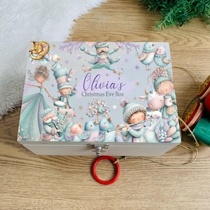 Puede incluir: Una caja rectangular de madera blanca con una colorida ilustración de personajes fantásticos, pájaros y un árbol. El texto "Olivia's Christmas Eve Box" está impreso en el centro. La caja tiene un cierre de metal plateado y un anillo rojo al frente.