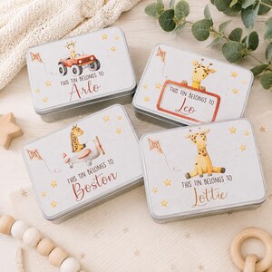 May include: Four rectangular metal tins featuring watercolour giraffe illustrations and personalised names Arlo, Leo, Boston, and Lottie. Each tin displays the text This tin belongs to followed by a name, decorated with small yellow stars and kites.