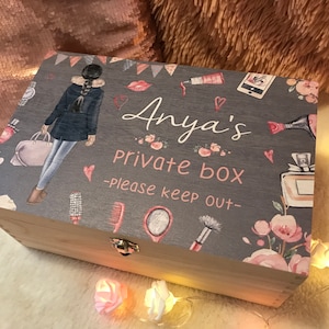 May include: A gray wooden box with a hinged lid. The lid is decorated with a colorful illustration of a girl, flowers, and makeup items. The text "Anya's Private Box - Please keep out -" is printed on the lid.